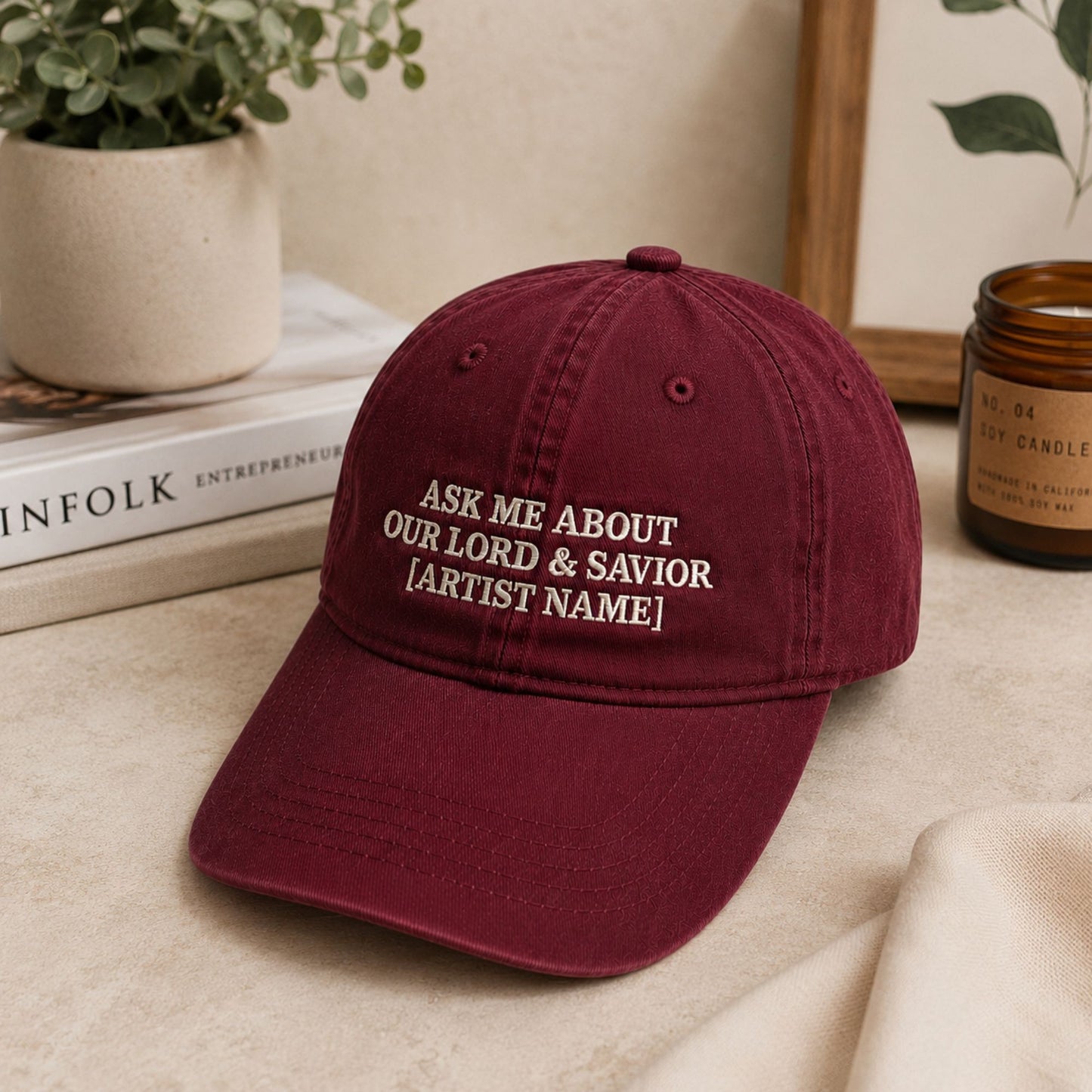 A maroon baseball cap with white embroidered text reading 'ASK ME ABOUT OUR LORD & SAVIOR [ARTIST NAME]', styled on a beige surface with books, a potted plant, and a candle in the background.