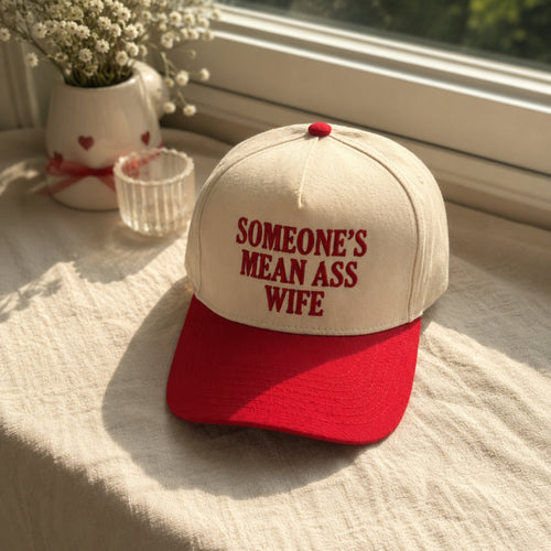 A baseball cap with an off-white crown and a red brim, featuring the text 'SOMEONE'S MEAN ASS WIFE' embroidered in red on the front. The cap is styled on a textured cream fabric surface next to a small glass cup and a white ceramic vase with dried flowers, with a window in the background.