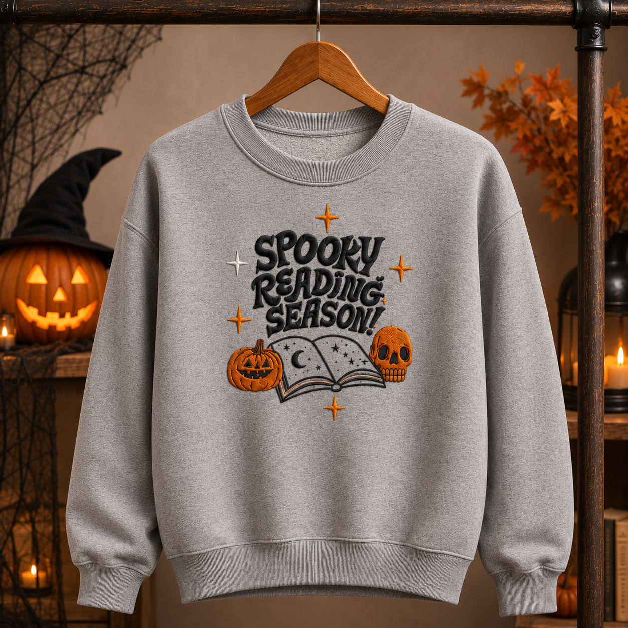 A heather gray crewneck sweatshirt with embroidered "Spooky Reading Season!" graphic featuring an open book, pumpkins, and a skull, displayed in a cozy fall Halloween setting.