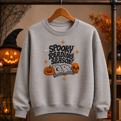 A heather gray crewneck sweatshirt with embroidered "Spooky Reading Season!" graphic featuring an open book, pumpkins, and a skull, displayed in a cozy fall Halloween setting.