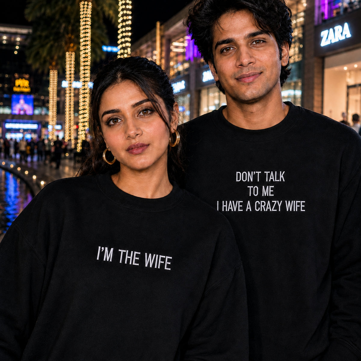 A man and a woman standing together at night wearing matching black crewneck sweatshirts with white text. The woman's sweatshirt reads 'I'M THE WIFE' and the man's reads 'DON'T TALK TO ME I HAVE A CRAZY WIFE'.