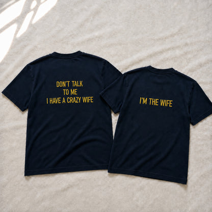 Two navy blue t-shirts shown from the back, laid flat on a light beige textured fabric background. The left shirt has yellow text reading 'DON'T TALK TO ME I HAVE A CRAZY WIFE' and the right shirt has yellow text reading 'I'M THE WIFE'.