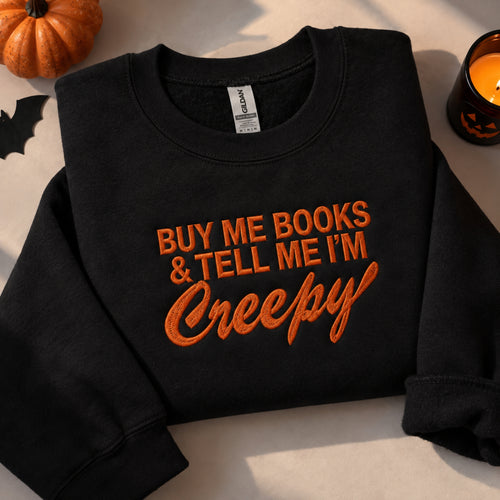 A black crewneck sweatshirt with orange embroidered text reading 'BUY ME BOOKS & TELL ME I'M Creepy' in a Halloween-themed flat lay, surrounded by a small pumpkin, a lit candle, and a black bat cutout.