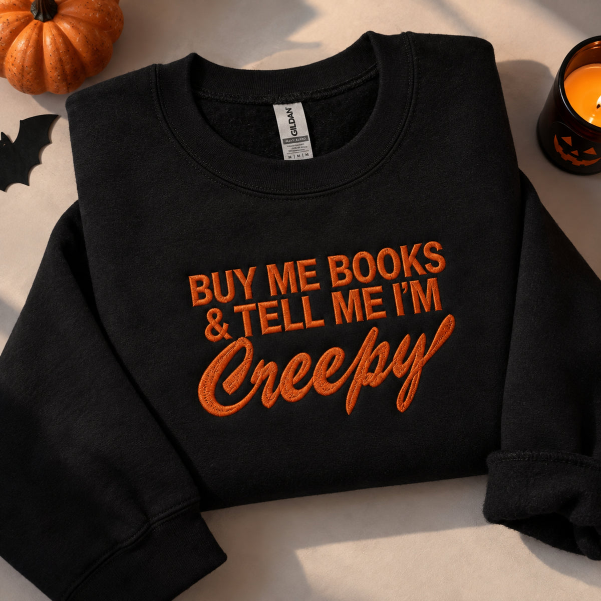 A black crewneck sweatshirt with orange embroidered text reading 'BUY ME BOOKS & TELL ME I'M Creepy' in a Halloween-themed flat lay, surrounded by a small pumpkin, a lit candle, and a black bat cutout.