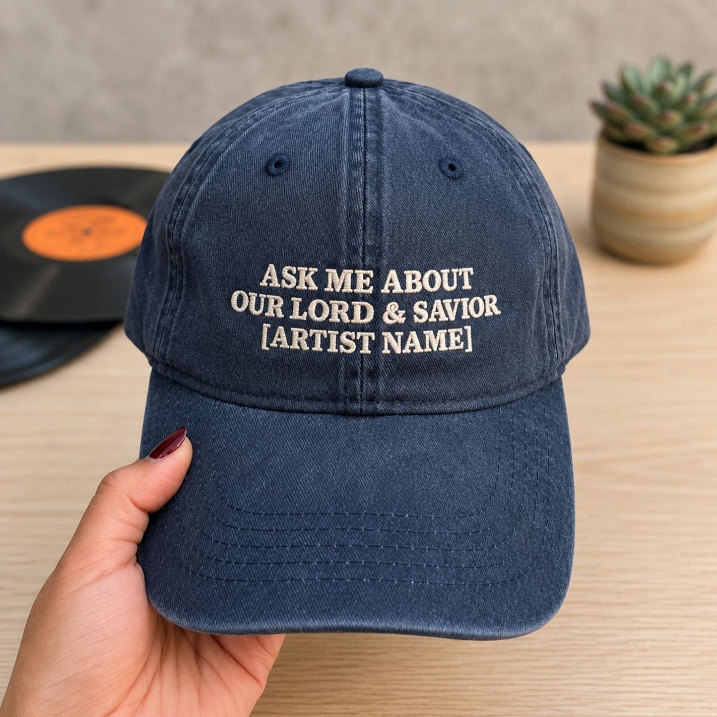 A navy blue baseball cap held by a hand, featuring white embroidered text that reads 'ASK ME ABOUT OUR LORD & SAVIOR [ARTIST NAME]'. The background shows a wooden table with a vinyl record and a small potted succulent.