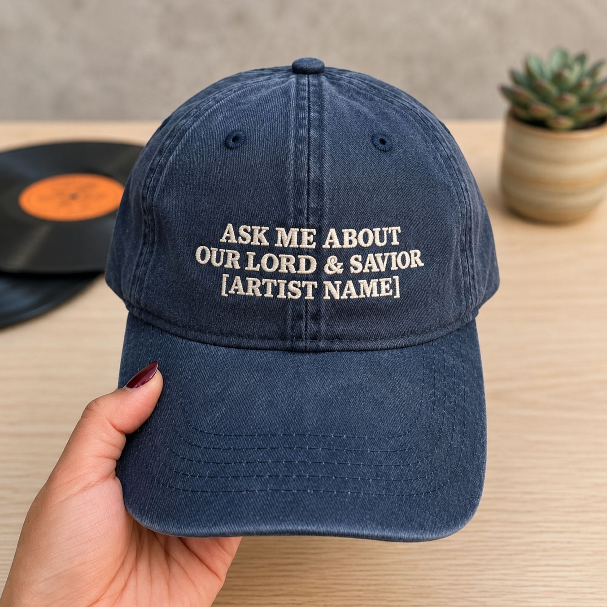 A navy blue baseball cap held by a hand, featuring white embroidered text that reads 'ASK ME ABOUT OUR LORD & SAVIOR [ARTIST NAME]'. The background shows a wooden table with a vinyl record and a small potted succulent.