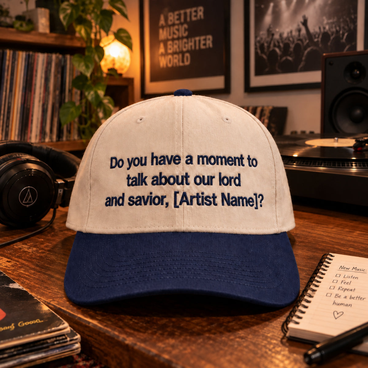 A baseball cap with a cream-colored crown and a navy blue brim, featuring the embroidered text 'Do you have a moment to talk about our lord and savior, [Artist Name]?'. The cap is displayed on a wooden desk surrounded by vinyl records, headphones, and a record player in a cozy music room setting.