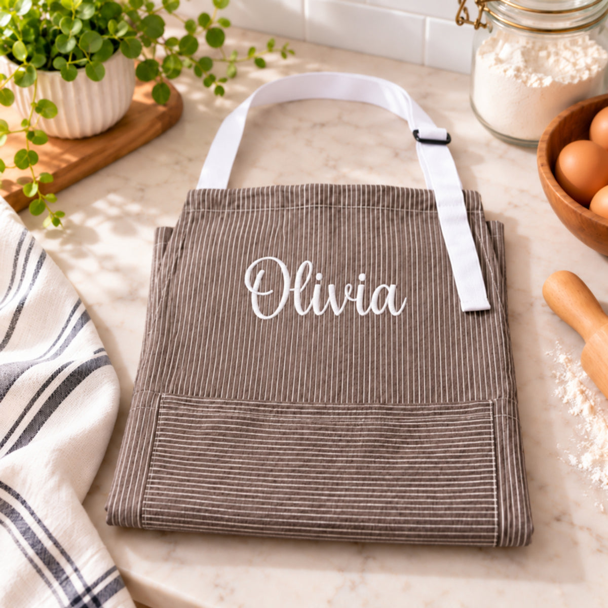 A brown corduroy kitchen apron with white striped handles and the name 'Olivia' embroidered in white script on the chest, displayed on a marble countertop with kitchen props.
