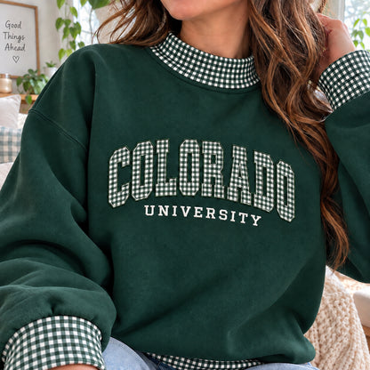 A woman wearing a dark green crewneck sweatshirt with a gingham check collar and cuffs, featuring 'COLORADO UNIVERSITY' printed in white block letters with a gingham pattern.