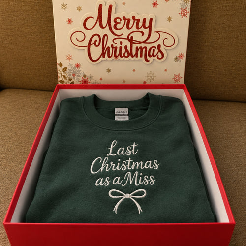 A folded dark green crewneck sweatshirt with white embroidered text reading 'Last Christmas as a Miss' and a bow graphic, presented inside a red gift box with a festive 'Merry Christmas' box lid behind it, set on a brown fabric surface.