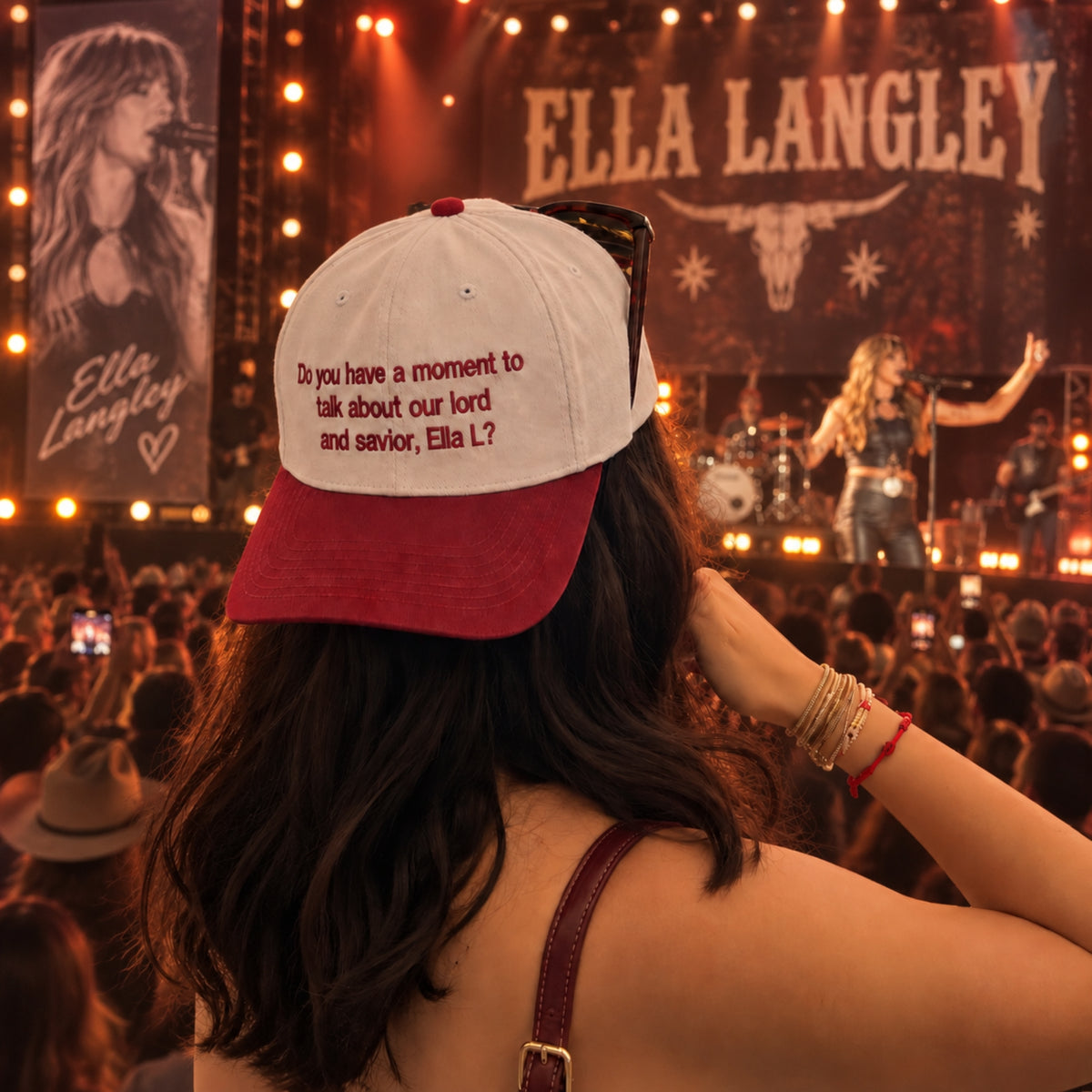 A woman at a live concert wearing a red and white baseball cap with the text 'Do you have a moment to talk about our lord and savior, Ella L?' and sunglasses resting on the cap. The background shows a brightly lit stage with 'ELLA LANGLEY' and a bull skull graphic.