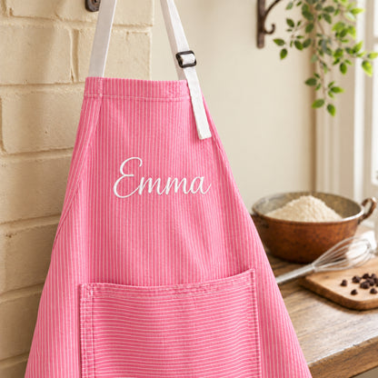 A pink striped kitchen apron with white straps and a front pocket, personalized with the name 'Emma' in white script. The apron is hanging on a wall hook in a warm, rustic kitchen setting with a wooden table, bowl of flour, and whisk in the background.