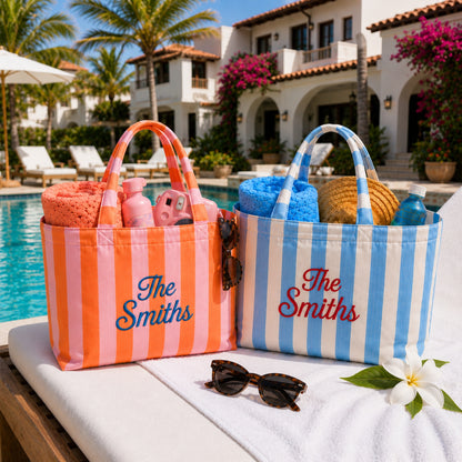 Two personalized beach tote bags with orange and pink stripes and blue and white stripes, filled with towels and beach accessories, sitting on a white lounge chair by a swimming pool with a resort building in the background.