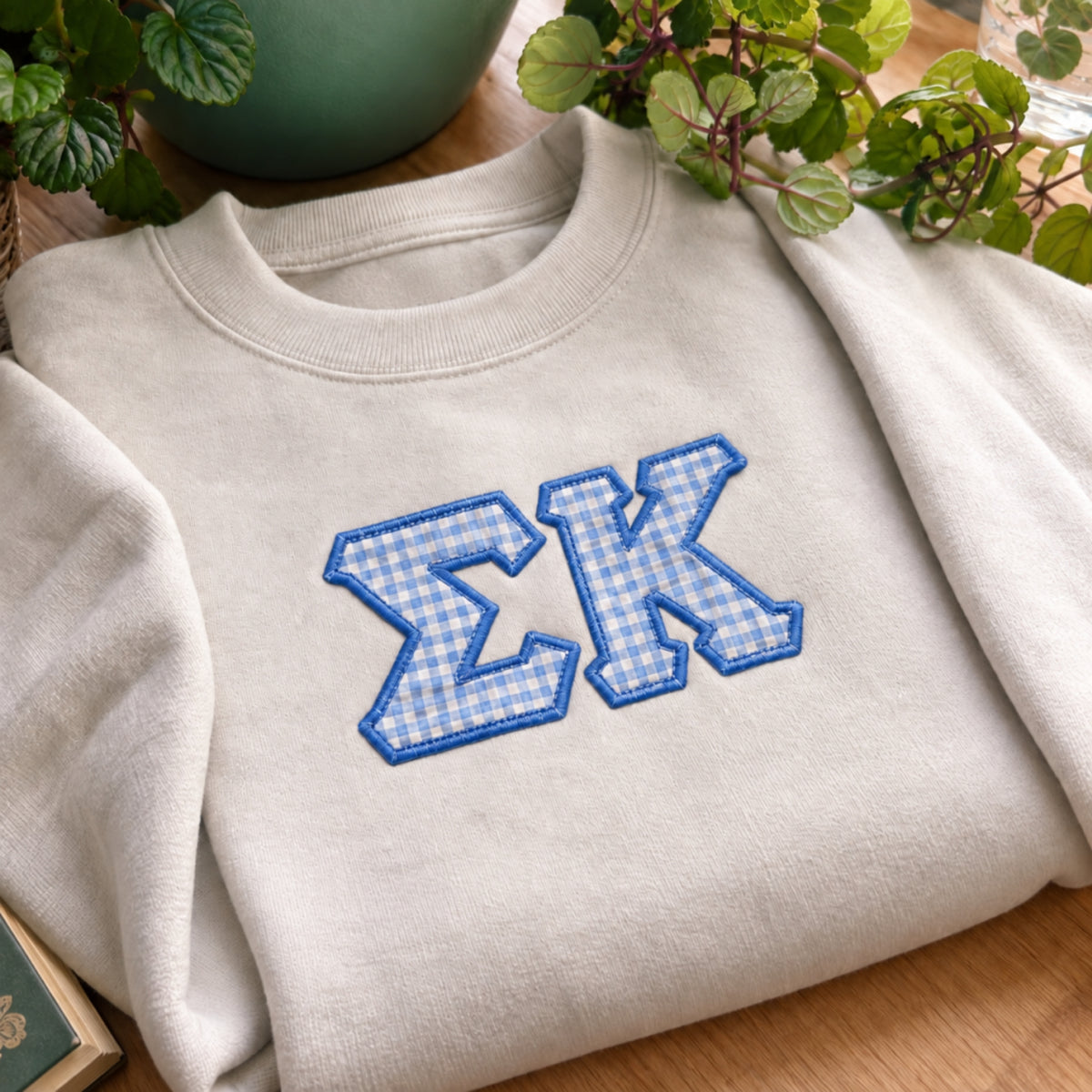A folded cream-colored crewneck sweatshirt with blue and white gingham embroidered Greek letters 'ΣΧ' on the chest, styled on a wooden surface with green plants in the background.