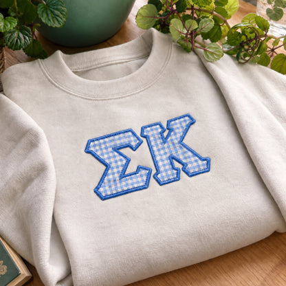 A folded cream-colored crewneck sweatshirt with blue and white gingham embroidered Greek letters 'ΣΧ' on the chest, styled on a wooden surface with green plants in the background.