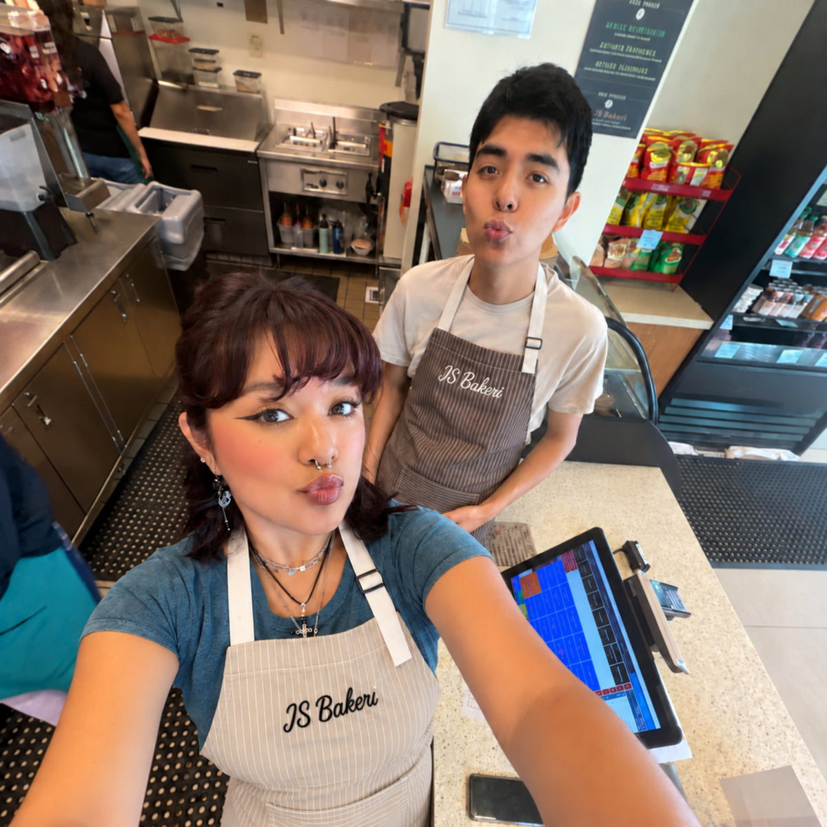 Two people wearing matching brown striped 'JS Bakers' aprons posing in a commercial kitchen setting.