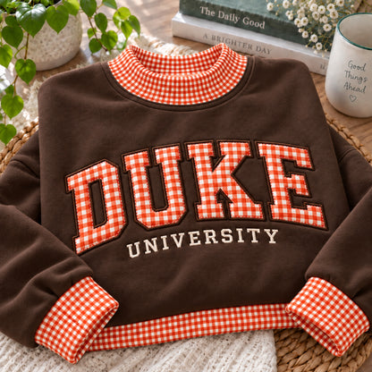 A brown sweatshirt with orange and white gingham trim on the collar, cuffs, and hem, featuring 'DUKE UNIVERSITY' embroidered across the front. The sweatshirt is styled in a cozy flat-lay setting with books, a mug, and a plant.