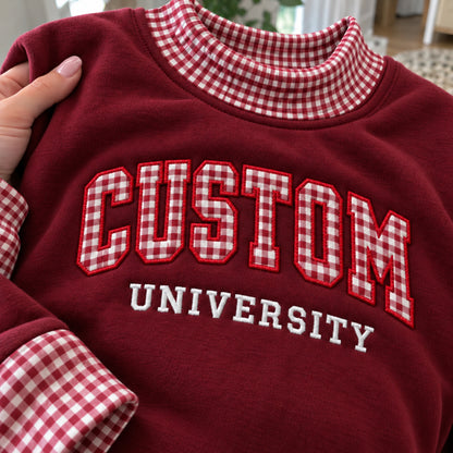 A close-up of a burgundy crewneck sweatshirt with red and white gingham check details on the collar and cuffs, featuring 'CUSTOM UNIVERSITY' embroidered on the front.