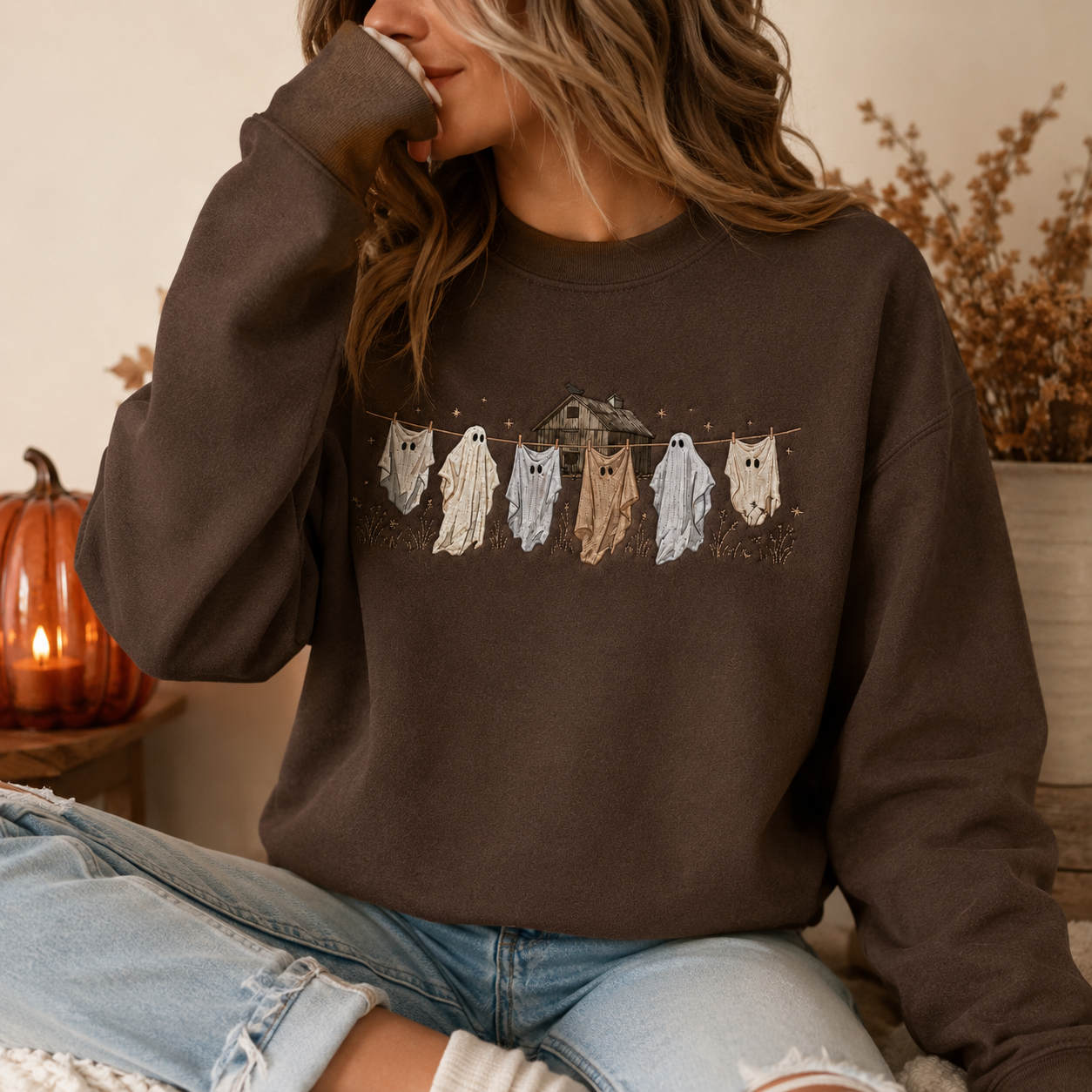 Woman wearing a dark brown Halloween crewneck sweatshirt featuring embroidered ghosts hanging on a clothesline with a haunted house design.