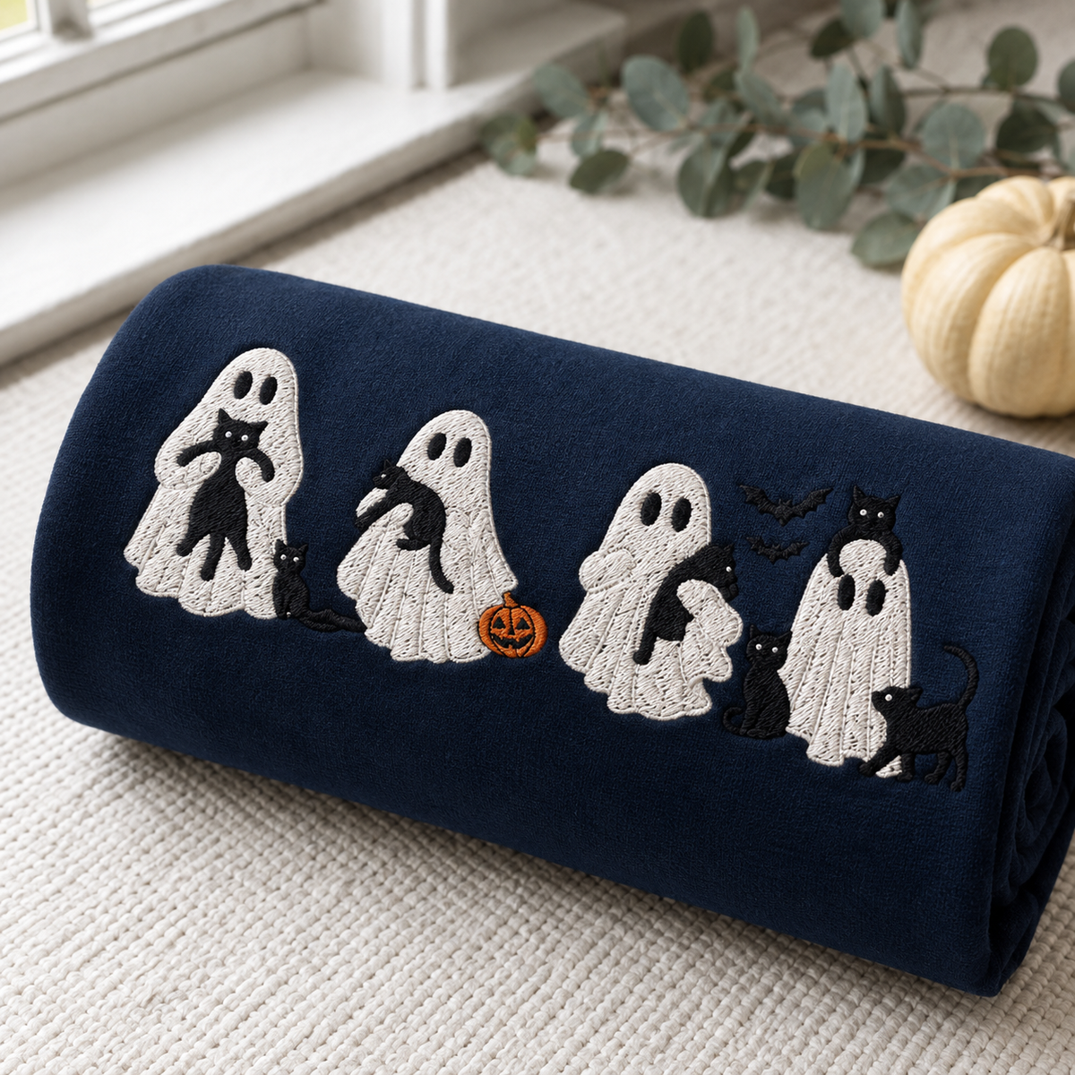 A navy blue blanket with embroidered Halloween ghosts, black cats, bats, and a small orange jack-o'-lantern, rolled up and styled on a textured cream surface with a small decorative pumpkin and eucalyptus leaves nearby.