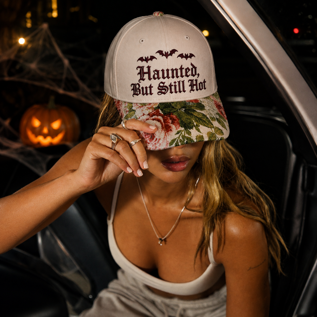 A woman sitting inside a car at night wearing a beige baseball cap with 'Haunted, But Still Hot' embroidered in gothic font and a floral brim. A glowing jack-o'-lantern and cobwebs are visible in the background.