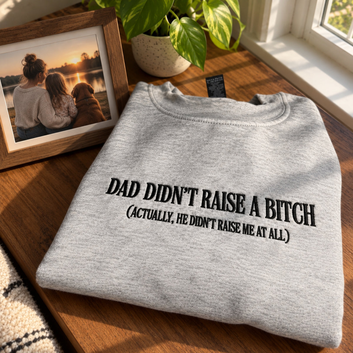 A folded light heather gray crewneck sweatshirt with black printed text reading 'DAD DIDN'T RAISE A BITCH (ACTUALLY, HE DIDN'T RAISE ME AT ALL)' sits on a wooden table next to a framed family photo and a potted plant.