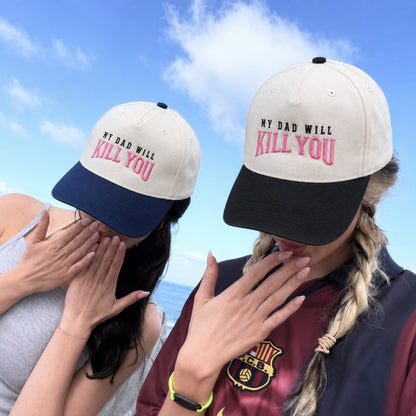 Two women outdoors under a blue sky wearing matching baseball caps with the text 'MY DAD WILL KILL YOU' in black and pink embroidery. The cap has an off-white crown, a dark navy brim, and a dark navy underside.