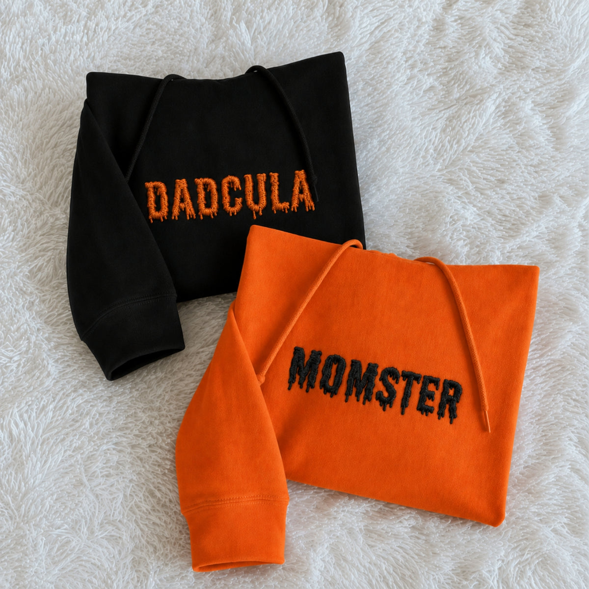 Two folded hooded sweatshirts laid flat on a white faux-fur surface: one black sweatshirt with orange 'DADCULA' text and one orange sweatshirt with black 'MOMSTER' text.