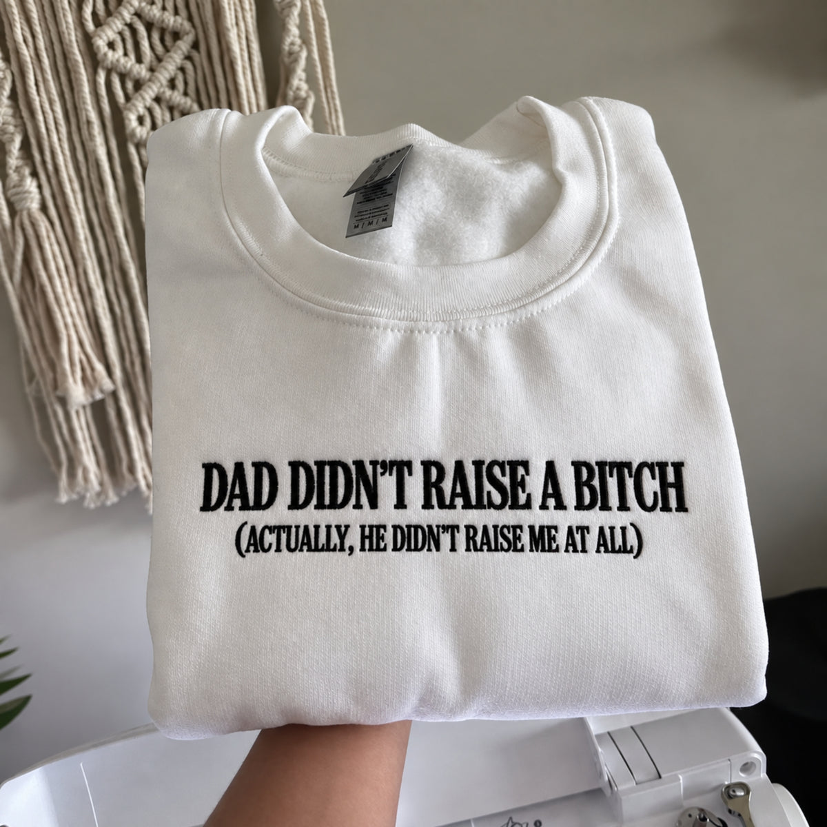 A folded white crewneck sweatshirt with black printed text reading 'DAD DIDN'T RAISE A BITCH (ACTUALLY, HE DIDN'T RAISE ME AT ALL)' held up by a hand in front of a neutral indoor background with a macrame wall hanging.
