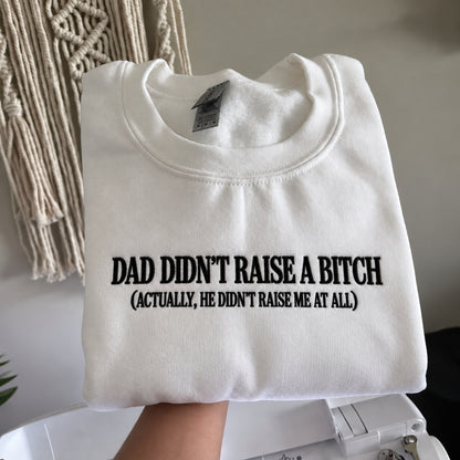 A folded white crewneck sweatshirt with black printed text reading 'DAD DIDN'T RAISE A BITCH (ACTUALLY, HE DIDN'T RAISE ME AT ALL)' held up by a hand in front of a neutral indoor background with a macrame wall hanging.