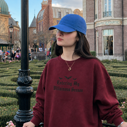 A woman wearing a blue baseball cap and a maroon crewneck sweatshirt with 'Entering My Villainous Season' printed in black gothic font, standing in front of a castle-themed background.