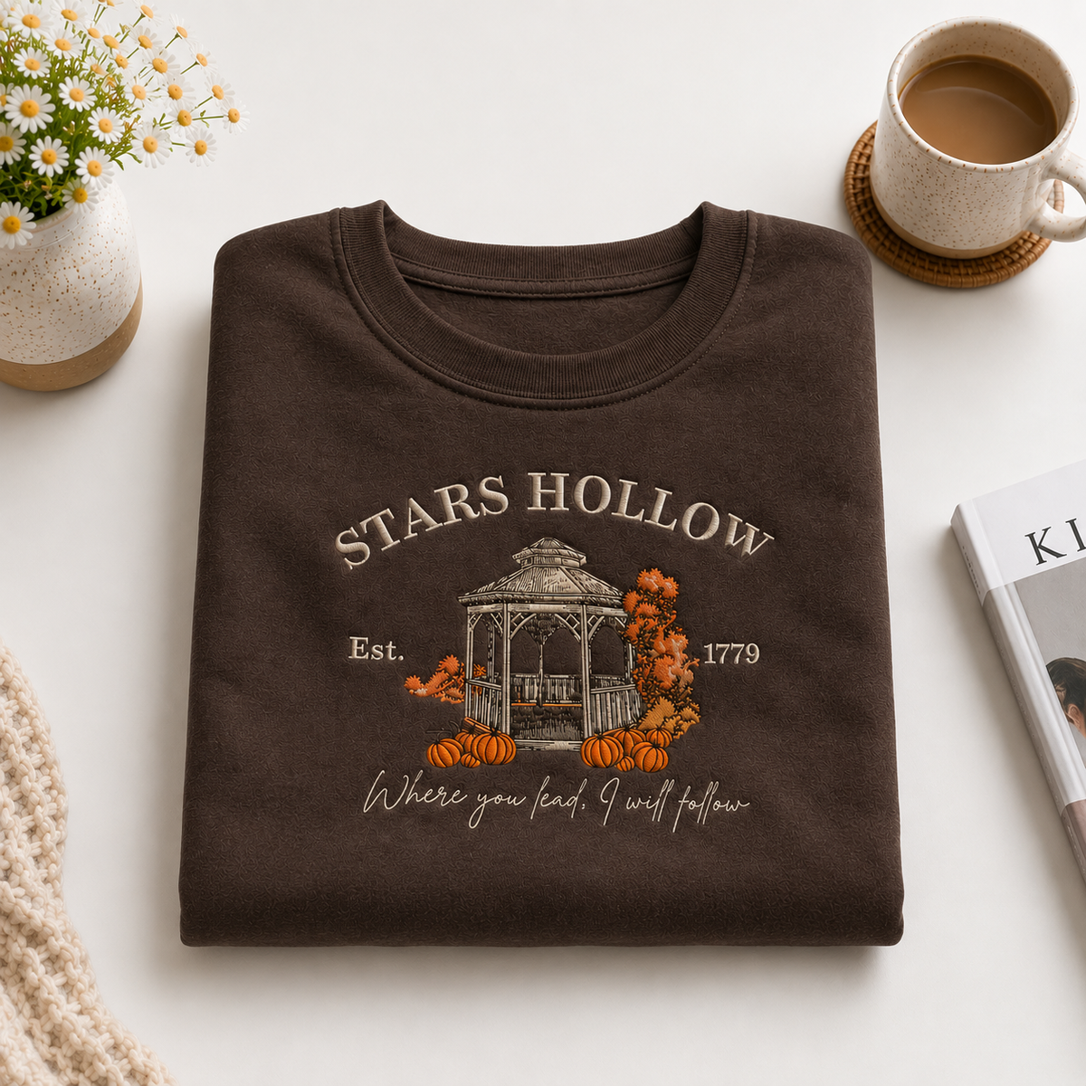 A folded dark brown graphic T-shirt is displayed on a light neutral surface with styled props, including a small flower vase, a cup of coffee, and a magazine. The shirt features a fall-themed graphic with the text 'STARS HOLLOW', 'Est.