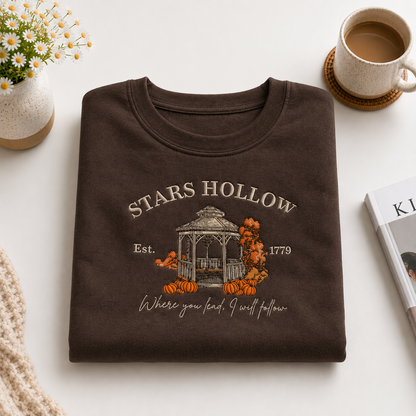 A folded dark brown graphic T-shirt is displayed on a light neutral surface with styled props, including a small flower vase, a cup of coffee, and a magazine. The shirt features a fall-themed graphic with the text 'STARS HOLLOW', 'Est.