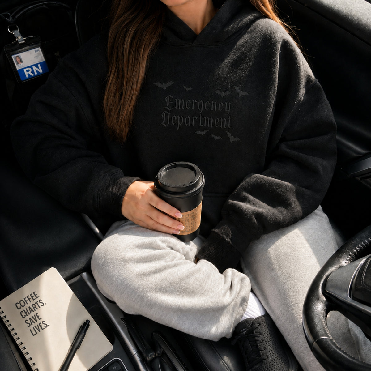 A person sitting in a car wearing a black hoodie with 'Emergency Department' printed in gothic font on the chest, holding a takeaway coffee cup.