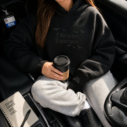 A person sitting in a car wearing a black hoodie with 'Emergency Department' printed in gothic font on the chest, holding a takeaway coffee cup.