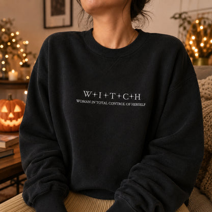 Woman wearing a black embroidered sweatshirt with the text “WITCH – Woman in Total Control of Herself” in a cozy Halloween-inspired setting.