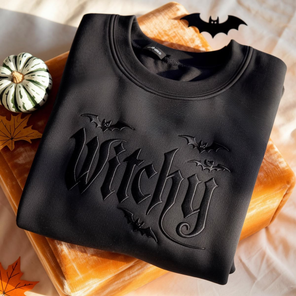A folded black crewneck sweatshirt with an embossed Gothic-style 'Witchy' graphic and bat motifs, styled on a stack of autumn-themed props including a small green-and-white pumpkin, orange leaves, and a wooden block.