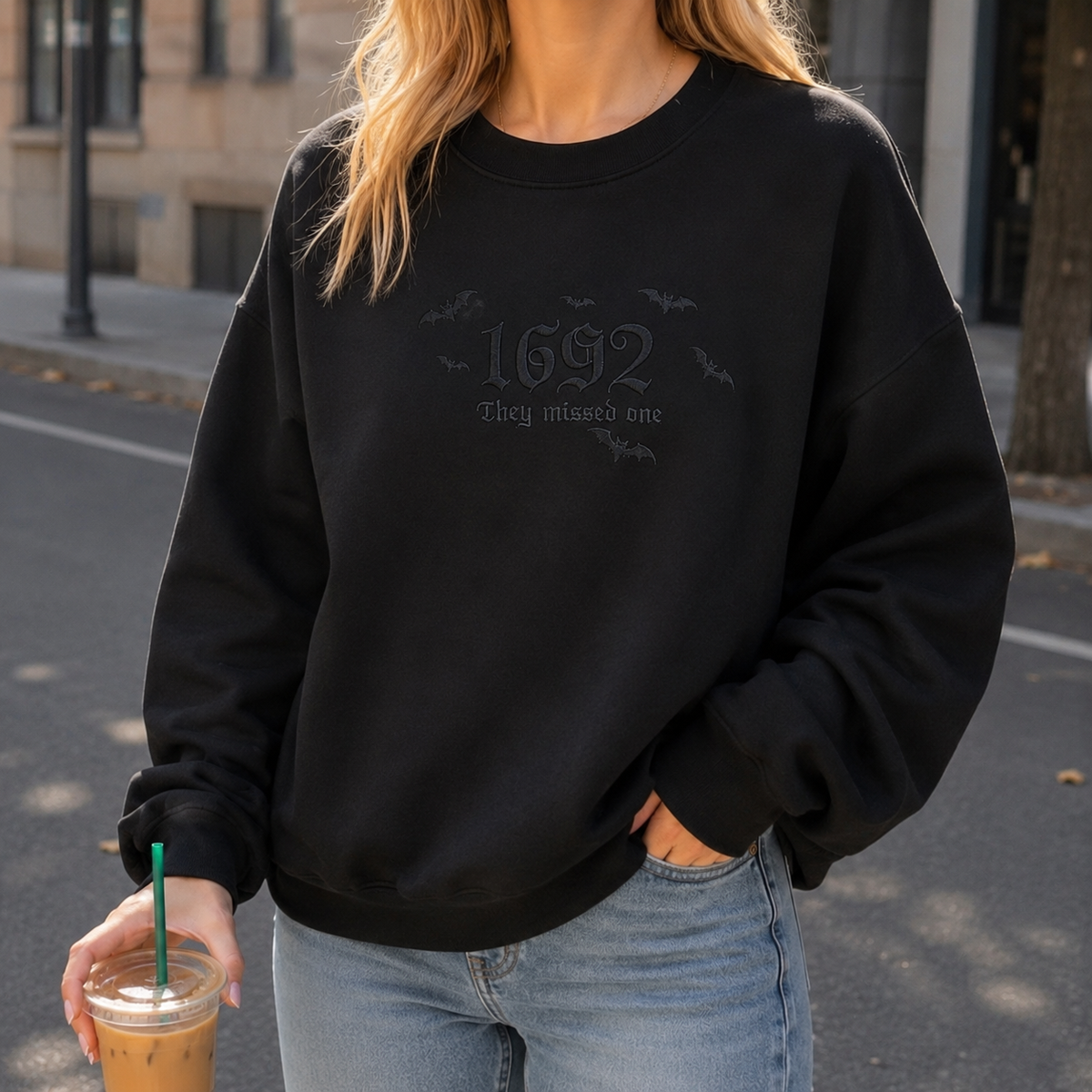 A woman wearing a black crewneck sweatshirt with a subtle tonal graphic on the chest that reads '1692 They missed one' with small bat silhouettes. She is also wearing light blue jeans and holding an iced coffee on a city street.