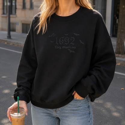 A woman wearing a black crewneck sweatshirt with a subtle tonal graphic on the chest that reads '1692 They missed one' with small bat silhouettes. She is also wearing light blue jeans and holding an iced coffee on a city street.