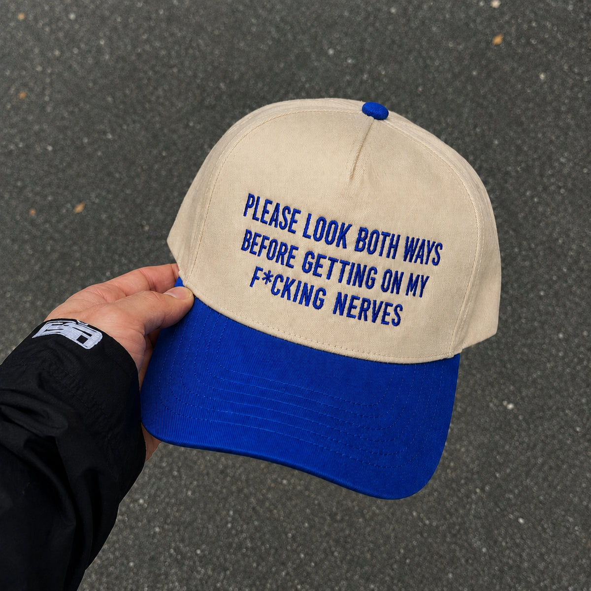 A beige and royal blue baseball cap held by a hand against an asphalt background. The front of the cap features blue embroidered text that reads, 'PLEASE LOOK BOTH WAYS BEFORE GETTING ON MY F*CKING NERVES'.