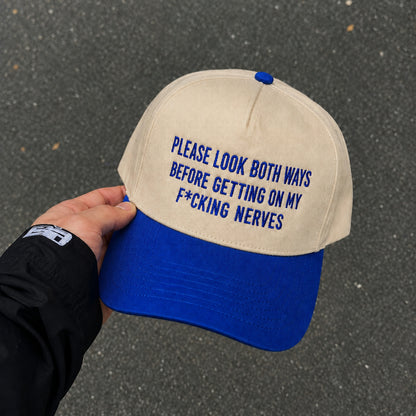 A beige and royal blue baseball cap held by a hand against an asphalt background. The front of the cap features blue embroidered text that reads, 'PLEASE LOOK BOTH WAYS BEFORE GETTING ON MY F*CKING NERVES'.