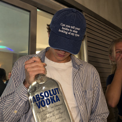 A person in a blue striped shirt and a navy blue baseball cap with the text 'Can you tell your boobs to stop looking at my eyes' holds a large bottle of Absolut Vodka. The photo is taken in a social setting with other people visible in the background.