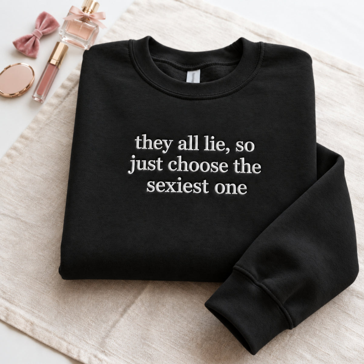 Folded black embroidered sweatshirt featuring the funny quote “they all lie, so just choose the sexiest one” in white stitching, displayed on a soft beige fabric background with pink perfume, makeup accessories, and feminine decor.