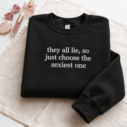 Folded black embroidered sweatshirt featuring the funny quote “they all lie, so just choose the sexiest one” in white stitching, displayed on a soft beige fabric background with pink perfume, makeup accessories, and feminine decor.