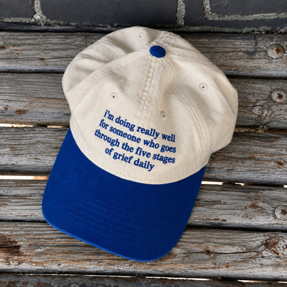 A baseball cap with a cream crown and a royal blue brim, featuring blue embroidered text that reads, 'I'm doing really well for someone who goes through the five stages of grief daily.' The cap is photographed from an angled top-down view on a rustic, weathered gray wooden plank surface.
