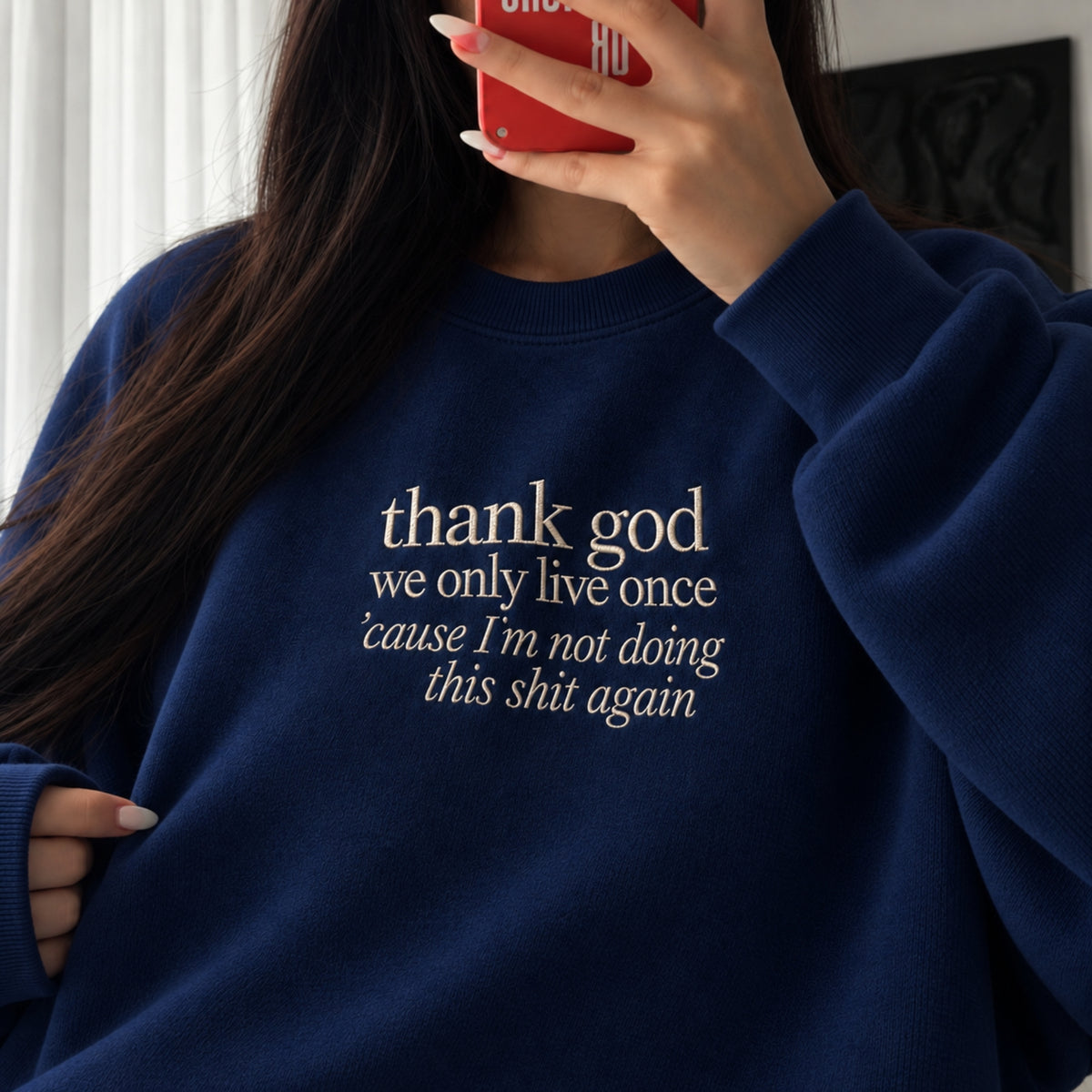 Woman wearing a navy blue embroidered sweatshirt with the white quote “thank god we only live once ’cause I’m not doing this shit again,” taking a mirror selfie indoors.