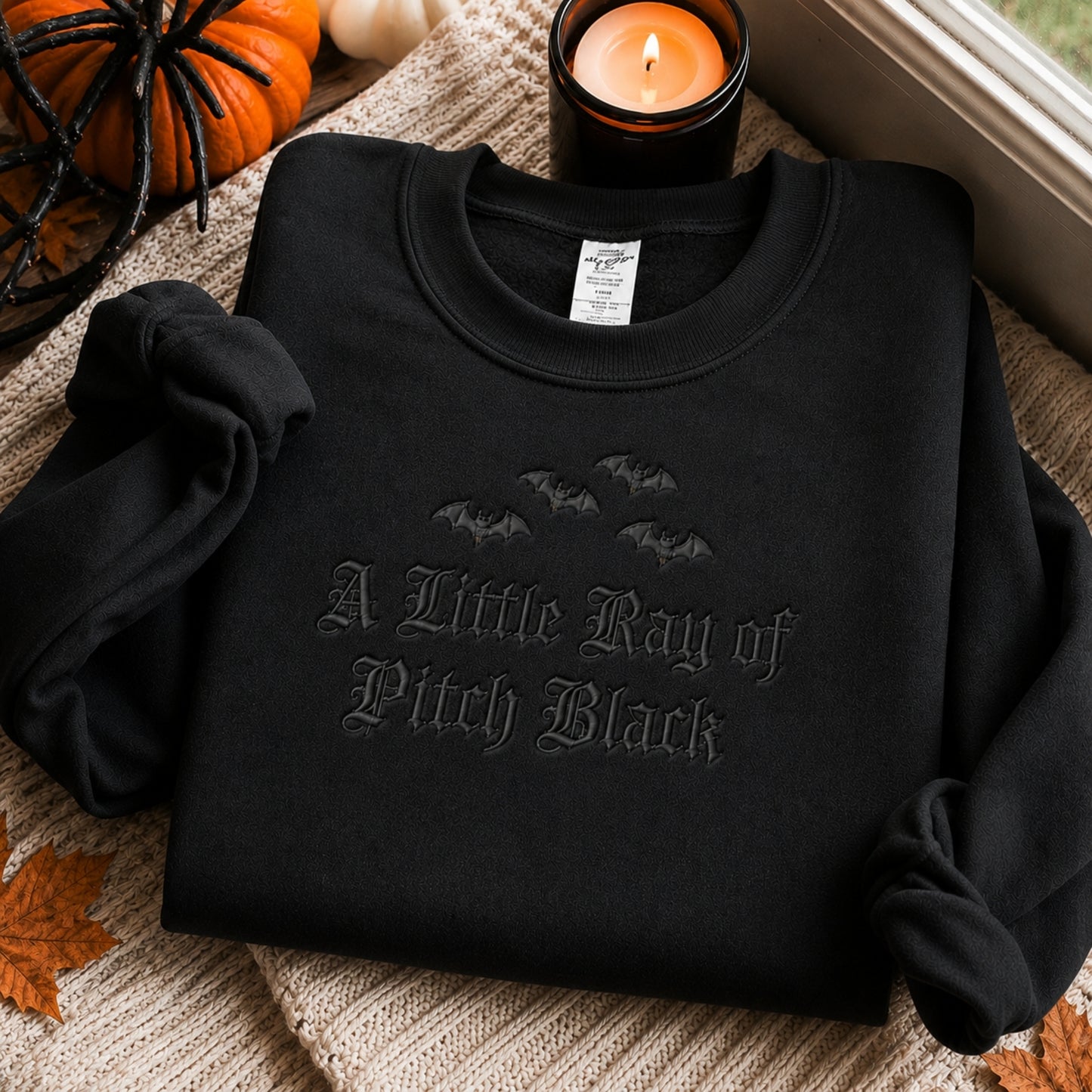 Folded dark humor embroidered sweater with premium black embroidery showcasing the A Little Ray of Pitch Black design, displayed beside candles and autumn decor for a cozy seasonal aesthetic.