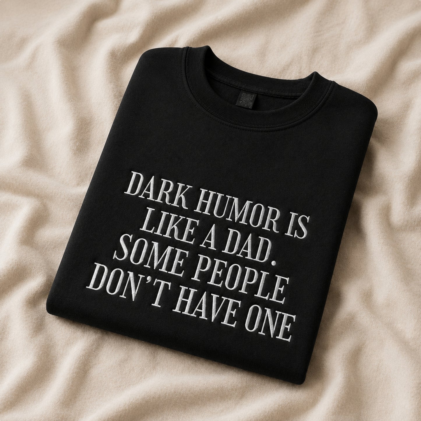 A folded black crewneck sweatshirt with white text reading 'DARK HUMOR IS LIKE A DAD. SOME PEOPLE DON'T HAVE ONE', displayed on a beige fabric background.