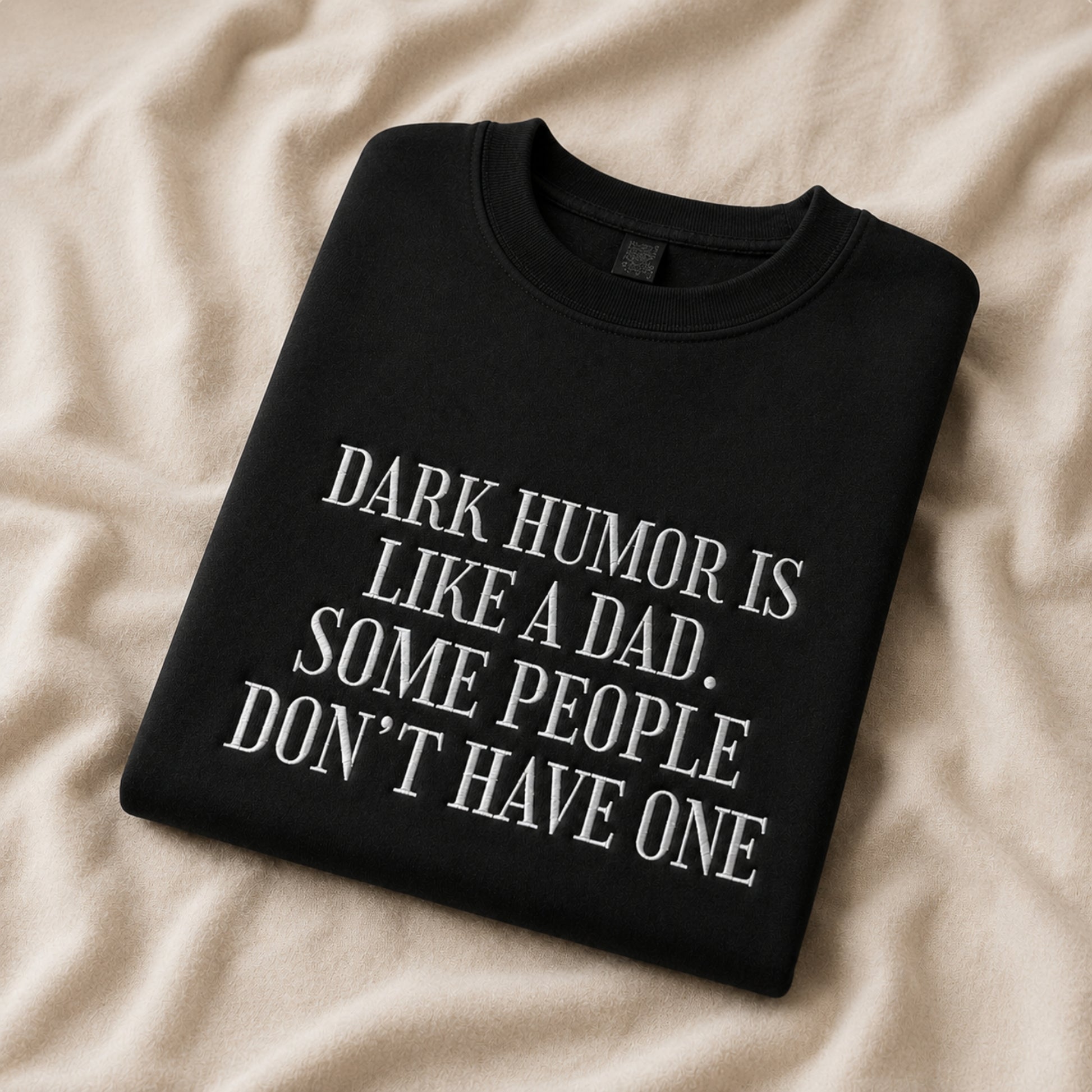 A folded black crewneck sweatshirt with white text reading 'DARK HUMOR IS LIKE A DAD. SOME PEOPLE DON'T HAVE ONE', displayed on a beige fabric background.