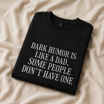 A folded black crewneck sweatshirt with white text reading 'DARK HUMOR IS LIKE A DAD. SOME PEOPLE DON'T HAVE ONE', displayed on a beige fabric background.